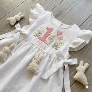 May include: A white sleeveless dress with ruffled sleeves and side ties, featuring embroidered daisies, a pink number one, a bunny, and the name Olivia. Three small, plush, cream-colored bunny-shaped toys are placed around the dress.