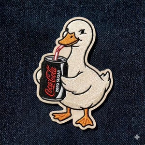 May include: Embroidered patch featuring a white duck holding a Coca-Cola Zero Sugar can with a red straw. The duck has an orange beak and feet. The Coca-Cola can is black with red lettering. The patch is on a dark blue denim background.