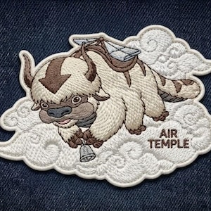 May include: An embroidered patch featuring a cream-colored, furry creature with brown markings, including an arrow on its head, flying through white clouds. The creature holds a small bell, and the text AIR TEMPLE is stitched in brown.