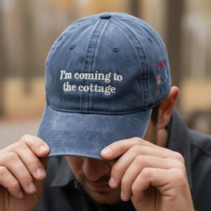 May include: A blue denim baseball cap with the embroidered text "I'm coming to the cottage" in white. The cap has a curved brim and a small embroidered design on the side.