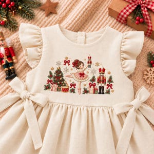 Peut inclure: Une robe couleur crème avec des manches à volants et des liens à la taille. Le corsage présente une scène brodée représentant une ballerine, un casse-noisette, un sapin de Noël, des cadeaux et des flocons dans des tons rouge, vert et brun.
