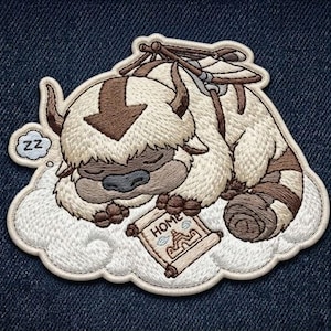May include: An embroidered patch featuring a sleeping Appa from Avatar: The Last Airbender, depicted with cream-colored fur, brown markings, and a dark arrow on the head. The creature rests on a white cloud, holding a scroll labeled HOME with a drawing.