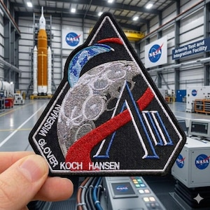 May include: A black, triangular NASA patch featuring a stylized moon, Earth, and red and blue accents. The patch includes the names WISEMAN, GLOVER, KOCH, and HANSEN. The background shows a rocket and the words "Artemis Test and Integration Facility."