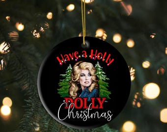 May include: A black ceramic Christmas ornament with the text "Have a Holly Dolly Christmas." The ornament features a portrait of a woman with blonde hair and a denim jacket, with two green Christmas trees on either side.