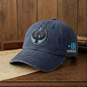 May include: Navy blue baseball cap with a circular patch featuring a black bird with outstretched wings and a star. The cap also has the text "IN STRONG" embroidered on the side. The hat is made of a soft, worn material.