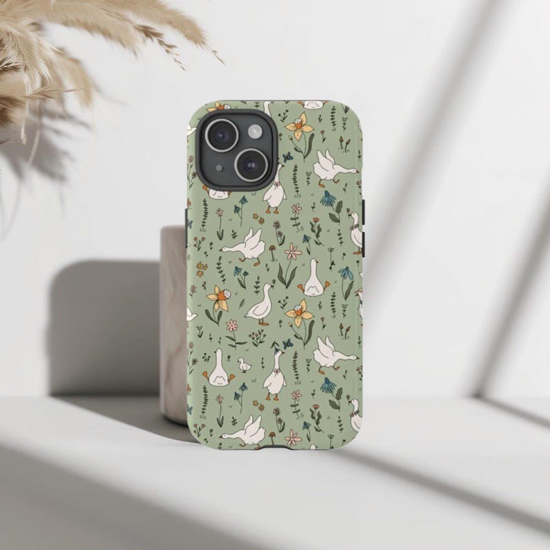 May include: A phone case with a whimsical pattern of white geese, yellow and blue flowers, and green foliage on a light green background. The case has a camera cutout and a smooth, glossy finish. The design is repeated across the entire surface.