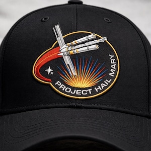 May include: A black baseball cap featuring an embroidered circular patch. The patch depicts a white spacecraft with a red trail, a yellow and blue sunburst, and the text PROJECT HAIL MARY in white capital letters against a black background.