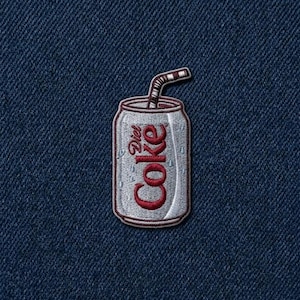 May include: An embroidered patch featuring a silver soda can with the red text Diet Coke and a striped straw, set against a dark blue denim fabric background. The design includes small light blue accents representing condensation droplets.
