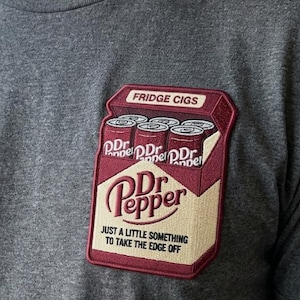May include: Dark gray t-shirt with a patch featuring a Dr. Pepper logo and the text "JUST A LITTLE SOMETHING TO TAKE THE EDGE OFF." The patch also includes the text "FRIDGE CIGS" and images of Dr. Pepper cans.