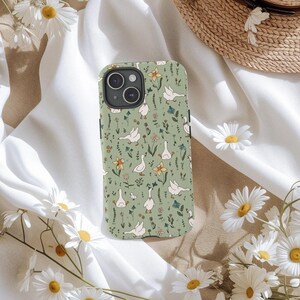 May include: A phone case with a green background featuring a pattern of white geese, yellow and blue flowers, and green foliage. The case has a camera cutout and black side buttons. Daisies and a straw hat are in the background.