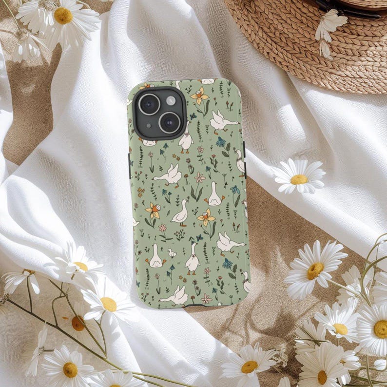 May include: A phone case with a green background featuring a pattern of white geese, yellow and blue flowers, and green foliage. The case has a camera cutout and black side buttons. Daisies and a straw hat are in the background.