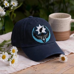 May include: A navy blue baseball cap featuring a circular embroidered patch on the front. The patch displays a black loon bird with outstretched wings centered against a light blue background and a white eight-pointed star shape behind it.