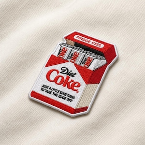 May include: Embroidered patch featuring a red and white Diet Coke cigarette pack design. The patch reads "Diet Coke" in a stylized font, with the text "JUST A LITTLE SOMETHING TO TAKE THE EDGE OFF." The top of the pack reads "FRIDGE CIGS."