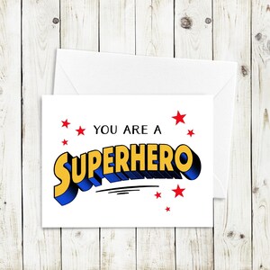 Puede incluir: Una tarjeta blanca con el texto "You are a Superhero" en una fuente de estilo cómic. El texto está en amarillo y azul con estrellas rojas alrededor.
