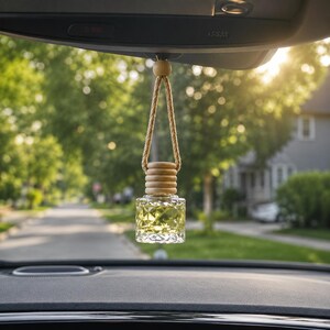 May include: A small, clear glass car air freshener bottle with a geometric, diamond-cut pattern filled with a yellow liquid. The bottle features a light-colored wooden cap and is suspended by a braided tan rope loop from a vehicle rearview mirror.