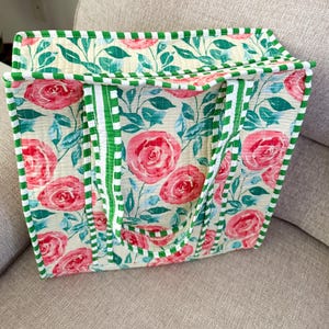 Pode incluir: Uma sacola floral com fundo creme, grandes rosas cor-de-rosa e folhas verdes. A sacola tem acabamento e alças listradas em verde e branco. A sacola está em uma superfície bege.