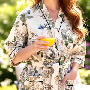 May include: A cream-colored floral robe with a green trim and a matching belt. The robe features a pattern of blue, green, and brown flowers. The robe has a relaxed fit and a wrap-around design.