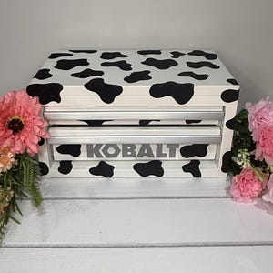 May include: A white and black cow print tool chest with two drawers. The word "KOBALT" is printed in gray on the front of the bottom drawer. Pink and peach flowers are on either side of the tool chest.