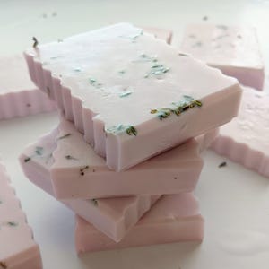 Puede incluir: Pila de jabones rectangulares de color rosa claro con borde festoneado. Los jabones tienen una superficie lisa con pequeñas motas verdes y doradas. Se ven otros jabones en el fondo. Cada jabón mide unos 8 cm de largo.
