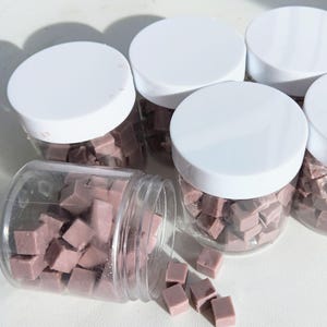 Puede incluir: Varios frascos de plástico transparente con tapas blancas, llenos de pequeños objetos cuadrados de color malva. Un frasco está volcado, derramando algunos de los cubos. Los frascos están colocados sobre una superficie blanca.