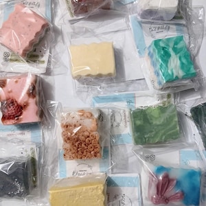 Könnte beinhalten: Sortiment handgemachter Seifen in verschiedenen Farben und Designs, einzeln in Klarsichtfolie verpackt. Die Seifen umfassen Rosa-, Gelb-, Grün-, Blau- und Brauntöne, wobei einige strukturierte Oberflächen und dekorative Elemente aufweisen. Jede Seife ist eine einzigartige Kreation.