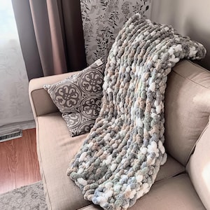 May include: A chunky knit throw blanket featuring a variegated color palette of soft white, sage green, light gray, and muted beige tones. The blanket is crafted from thick, soft yarn loops, creating a highly textured, plush, and voluminous appearance.
