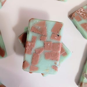 May include: Several rectangular bars of soap with a light blue base and a mosaic-like pattern of reddish-brown pieces. The soap bars are arranged on a white surface, with some overlapping. The overall aesthetic is clean and minimalist.