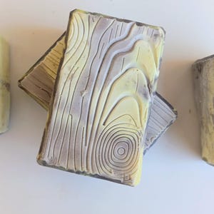 May include: Four rectangular bars of soap with a wood grain design. The soap has a yellow and purple color scheme. Two bars are stacked, and two are standing upright. The soap has a textured appearance.