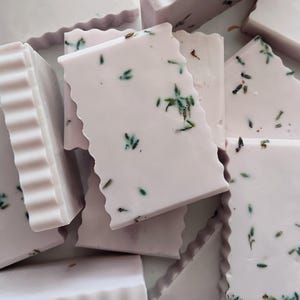 Puede incluir: Primer plano de varias pastillas de jabón de color lavanda con un borde ondulado. Las pastillas de jabón son rectangulares y tienen pequeñas motas verdes y marrones. Las pastillas de jabón están apiladas unas sobre otras.