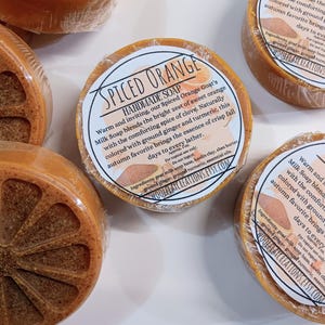 Puede incluir: Barras redondas de jabón artesanal de leche de cabra Spiced Orange envueltas en plástico transparente. Cada barra presenta una superficie texturizada de color naranja con un patrón de rodaja de cítrico y una etiqueta de papel circular.
