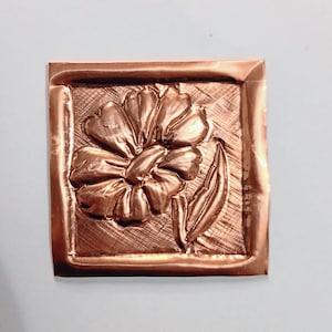 May include: A square copper relief sculpture featuring a detailed flower with multiple petals and a single leaf. The flower is raised from the surface, creating a three-dimensional effect. The copper has a warm, reddish-brown hue.