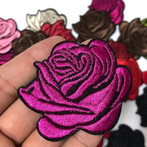 May include: A collection of embroidered rose patches in shades of pink, red, brown, and black. The image highlights a magenta rose patch with black detailing. These patches are suitable for fabric decoration.