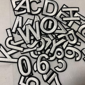 May include: A collection of white embroidered letters and numbers with black borders. The assortment includes various letters of the alphabet and numbers, suitable for crafting or personalization projects. The patches are scattered on a light-colored surface.