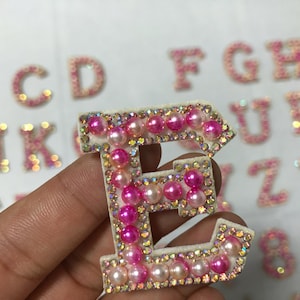 May include: Close-up of a sparkling letter "E" with pink pearls and iridescent rhinestones. The letter is held in a hand, with other glittery letters and numbers in the background. The letters are likely for crafts or embellishments.