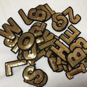 May include: A collection of gold sequined letters and numbers with black borders. The alphabet and numbers are scattered across a white surface. The sequins give a shiny, textured appearance to the patches.