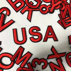 Può includere: Lettere e numeri rossi ricamati su sfondo bianco. La parola "USA" è ben visibile. Altre lettere e numeri sono sparsi. Le lettere e i numeri hanno un contorno nero.