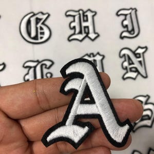 Può includere: Toppa ricamata bianca con la lettera "A" e bordo nero, tenuta in mano. La toppa è in un carattere inglese antico. Altre lettere dello stesso stile sono visibili sullo sfondo.