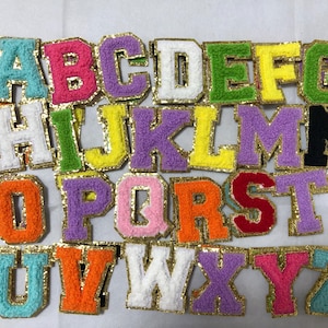 May include: A collection of colorful, embroidered fabric letters, each outlined in gold glitter. The letters are in various colors, including teal, pink, purple, and yellow, and are arranged on a white surface. The letters are for crafts and personalization.