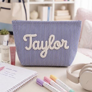 Può includere: Una pochette con cerniera a righe blu e bianche con il nome Taylor in rilievo realizzato con corda bianca sul davanti. La pochette è rettangolare con la parte superiore affusolata ed è posizionata tra vari articoli di cancelleria e prodotti.