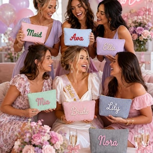 May include: A collection of rectangular zippered pouches featuring vertical pinstripes in various colors, each embroidered with a unique name or title including Maid, Ava, Ella, Mia, Bride, Lily, and Nora in contrasting script lettering.