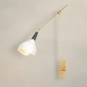 Op de afbeelding: Een moderne wandlamp met een witte, klokvormige kap en een verstelbare arm in messingkleur. De lamp heeft een zwart accent aan de basis van de kap en een rechthoekige messing montageplaat. De arm van de lamp is ongeveer 50 cm lang.