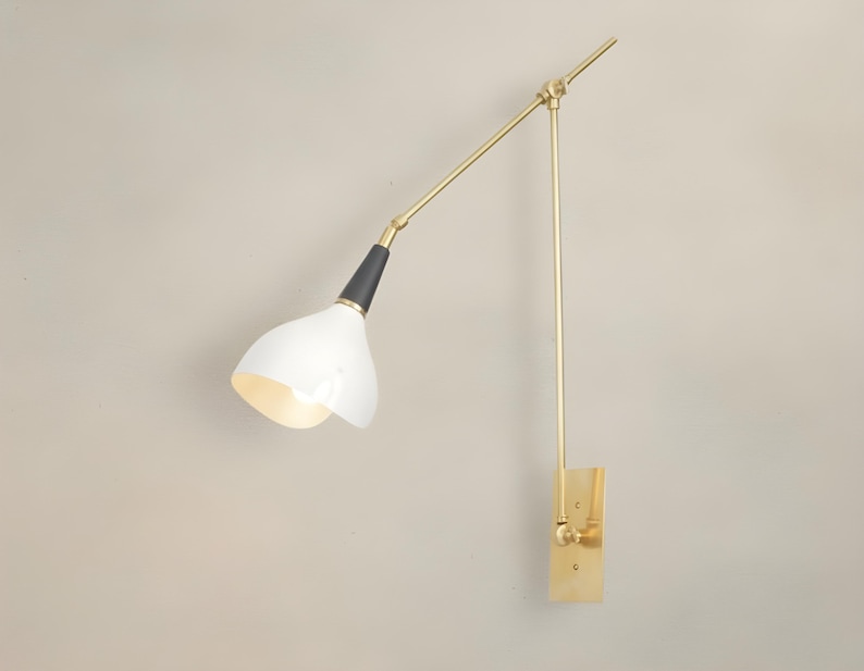 Op de afbeelding: Een moderne wandlamp met een witte, klokvormige kap en een verstelbare arm in messingkleur. De lamp heeft een zwart accent aan de basis van de kap en een rechthoekige messing montageplaat. De arm van de lamp is ongeveer 50 cm lang.