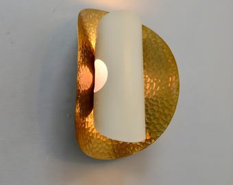Pode incluir: Arandela de parede com uma parte traseira de metal texturizado dourado. Uma vela cilíndrica branca é colocada no centro, com uma pequena abertura que revela uma luz quente e brilhante. A arandela é montada numa parede cinza claro.