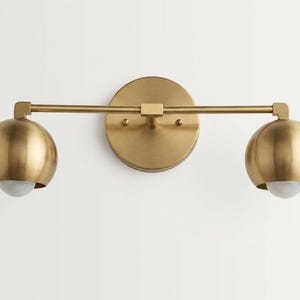 Pode incluir: Uma luminária de parede com uma barra horizontal de latão fixada a uma placa circular. Duas cúpulas esféricas de latão estão posicionadas em cada extremidade da barra, cada uma contendo uma pequena lâmpada de vidro fosco branco.