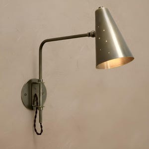 May include: A wall-mounted, silver-toned lamp with a conical shade featuring small, evenly spaced perforations. The lamp has a curved arm and a circular base. The shade is angled downward, and the interior is a warm, golden color.