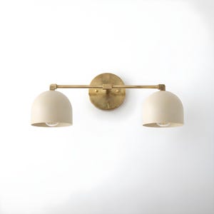 May include: A two-light vanity light fitting with a brass finish. It features two off-white, dome-shaped shades. The shades are attached to a horizontal bar extending from a circular backplate.