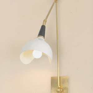 Op de afbeelding: Een wandlamp met een witte, gebogen kap en een zwart-gouden arm. De lamp heeft een rechthoekige basis in messingkleur en een lange, verstelbare arm. De gloeilamp is zichtbaar in de kap. De lamp is ontworpen voor gebruik binnenshuis.