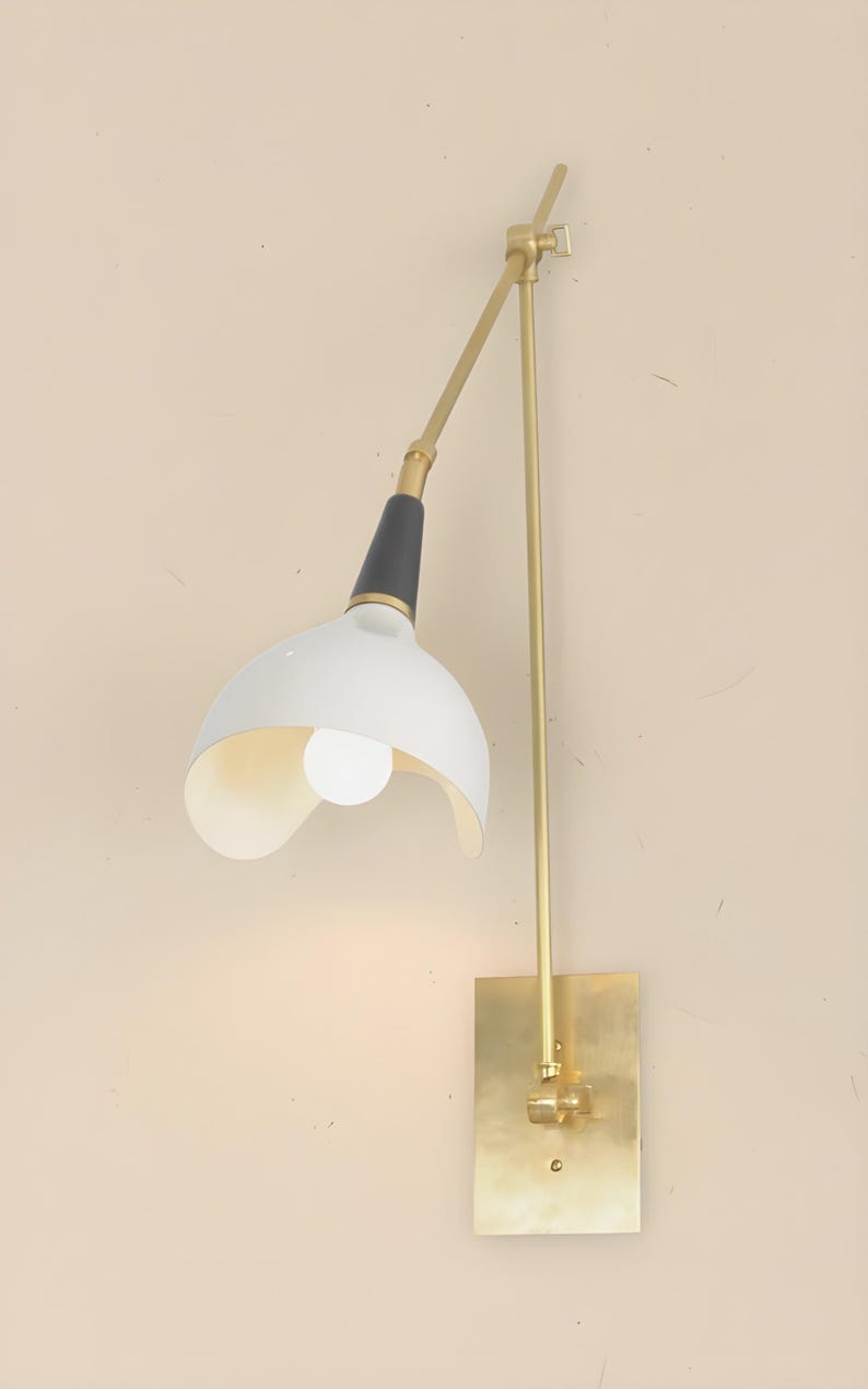 Op de afbeelding: Een wandlamp met een witte, gebogen kap en een zwart-gouden arm. De lamp heeft een rechthoekige basis in messingkleur en een lange, verstelbare arm. De gloeilamp is zichtbaar in de kap. De lamp is ontworpen voor gebruik binnenshuis.