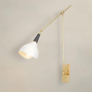 Op de afbeelding: Een moderne wandlamp met een witte, klokvormige kap en een verstelbare arm in messingkleur. De lamp heeft een zwart accent aan de basis van de kap en een rechthoekige messing montageplaat. De arm van de lamp is ongeveer 50 cm lang.