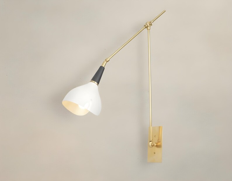 Op de afbeelding: Een moderne wandlamp met een witte, klokvormige kap en een verstelbare arm in messingkleur. De lamp heeft een zwart accent aan de basis van de kap en een rechthoekige messing montageplaat. De arm van de lamp is ongeveer 50 cm lang.